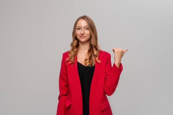 Portrait of smiling confident blonde young woman pointing finger to the right, showing advertisement, looking determined at camera, standing in red blazer against gray background - Photo, Image