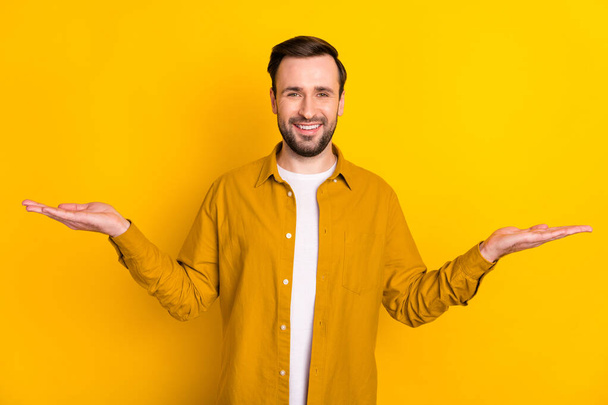 Photo of young man happy positive smile hold hands product promo offer pros cons ad isolated over yellow color background. - Fotografie, Obrázek