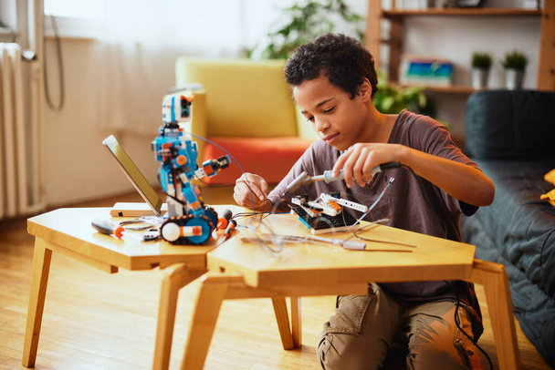 Ένα Αφροαμερικανάκι που φτιάχνει ρομπότ στο σπίτι. Εκπαίδευση στη ρομποτική στο σπίτι. - Φωτογραφία, εικόνα