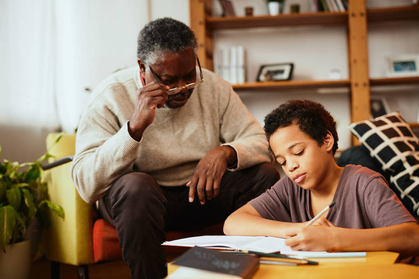 An African-American grandfather is sitting at home with his grandson and helping him with homework. - Фото, зображення