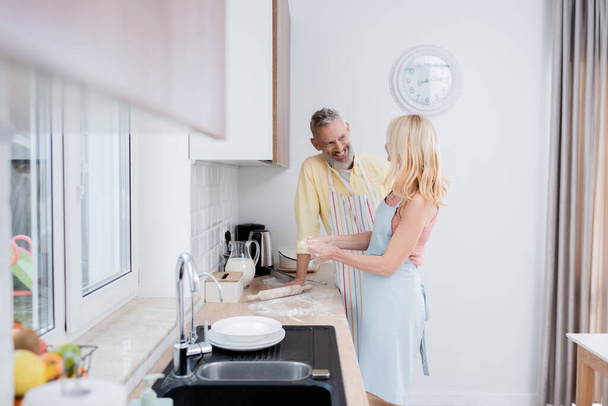 Усміхнений чоловік дивиться на дружину, роблячи тісто біля борошна і прокатного штифта на кухні
  - Фото, зображення