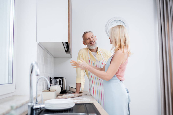 Positive mature man talking to wife with dough in kitchen  - Фото, зображення