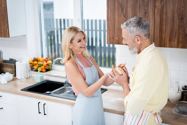Smiling blonde woman in apron holding dough near husband in kitchen  - Fotografie, Obrázek