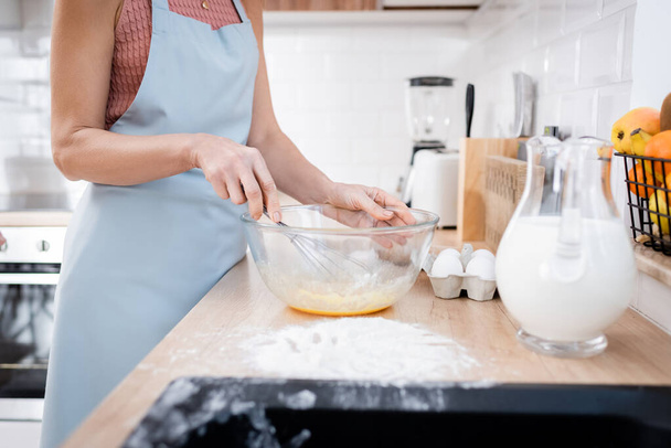 Vista ritagliata della donna in grembiule mescolando le uova nella ciotola vicino alla farina e al latte sul piano di lavoro della cucina  - Foto, immagini