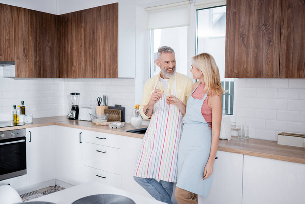 Smiling mature couple in aprons holding champagne in kitchen  - Фото, изображение