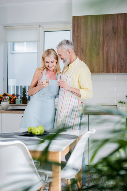 Позитивный зрелый мужчина в фартуке держит шампанское рядом с женой в фартуке на кухне  - Фото, изображение