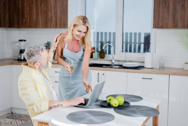 Femme souriante dans le tablier tenant champagne près du mari en utilisant un ordinateur portable dans la cuisine  - Photo, image