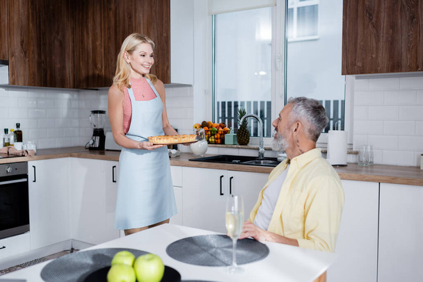 Femme positive tenant tarte maison près du mari et champagne sur la table dans la cuisine  - Photo, image