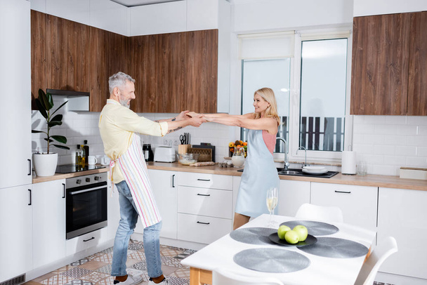 Pozitivní pár v zástěrách tančí u šampaňského na stole v kuchyni  - Fotografie, Obrázek