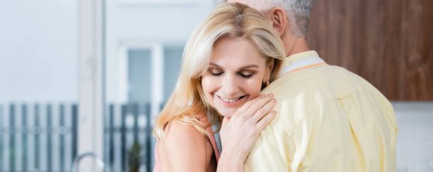Mature woman smiling while hugging husband in kitchen, banner  - 写真・画像