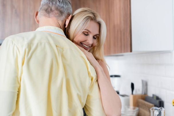 Smiling woman hugging mature husband in kitchen  - Fotó, kép