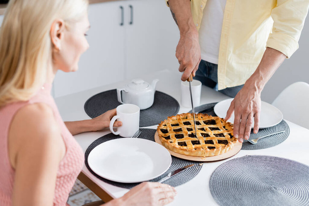 Mature man cutting pie near blurred wife with tea at home  - Foto, Bild