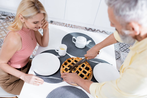 Overhead view of blurred man cutting homemade pie near smiling wife in kitchen  - Foto, imagen