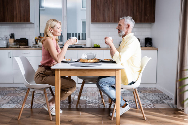 Side view of positive mature couple holding cups near pie in kitchen  - Foto, Imagem