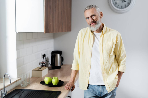Lächelnder älterer Mann blickt in Küche in die Kamera neben Äpfeln  - Foto, Bild