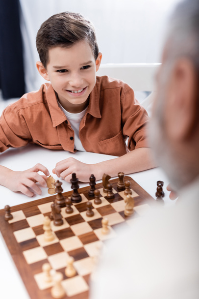 cheerful boy playing chess with blurred grandpa at home - Фото, изображение