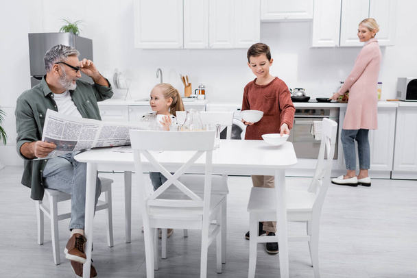smiling man with newspaper looking at grandchildren setting table for breakfast - Photo, Image