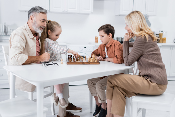 girl holding figure while playing chess with grandparents and brother in kitchen - Photo, Image