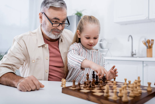 Ένας ηλικιωμένος με γυαλιά χαμογελά κοντά στην εγγονή του παίζοντας σκάκι. - Φωτογραφία, εικόνα