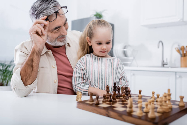 happy senior man holding eyeglasses while playing chess with little granddaughter in kitchen - Fotó, kép