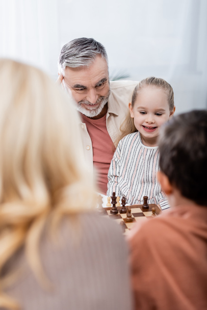 ανώτερος άνθρωπος χαμογελά ενώ παίζει σκάκι με τα εγγόνια και τη σύζυγο, θολή πρώτο πλάνο - Φωτογραφία, εικόνα