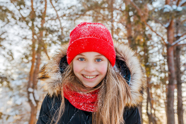 Portrait of a charming little girl in a red hat in a snowy forest in winter. Christmas winter holidays. Happy childhood - Photo, image