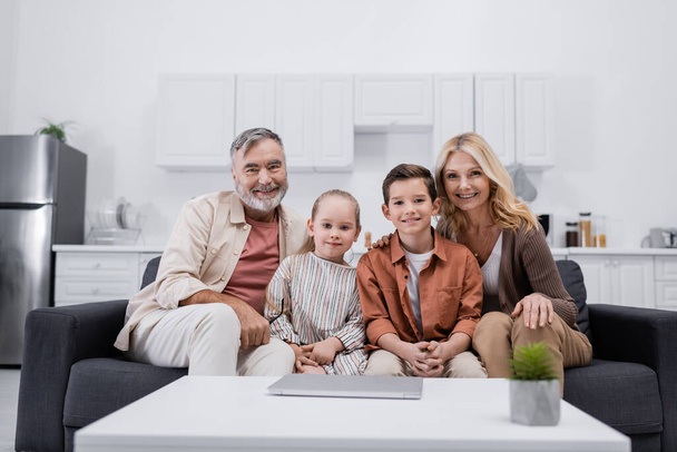 happy couple and grandchildren sitting on couch in kitchen and looking at camera - Fotoğraf, Görsel