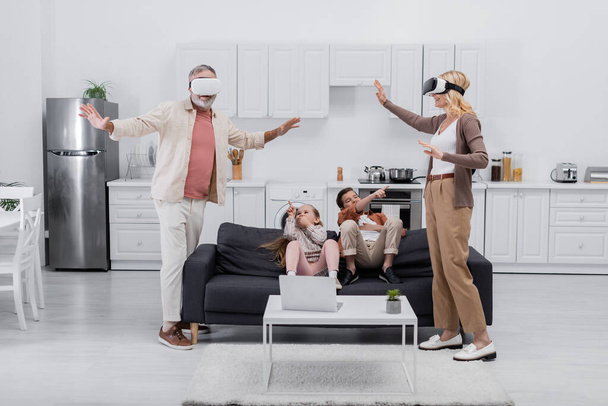 kids pointing with fingers at grandparents gaming in vr headsets in kitchen - Foto, Imagen