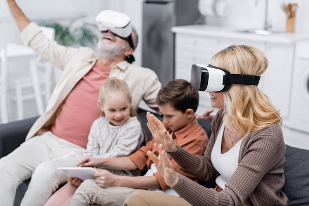 excited couple gaming in vr headsets near grandchildren using digital tablet on couch - Valokuva, kuva