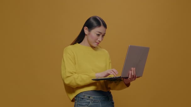 Aasialainen nainen käyttää kannettavaa tietokonetta kameran edessä keltaisella taustalla - Materiaali, video