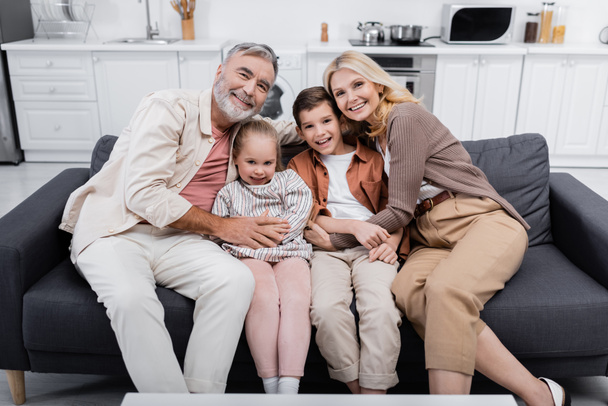 cheerful couple hugging grandchildren and looking at camera on sofa at home - Fotografie, Obrázek