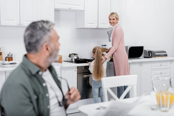 hymyilevä nainen katselee hämärtynyt aviomies valmistellessaan aamiaista tyttärentyttären kanssa - Valokuva, kuva