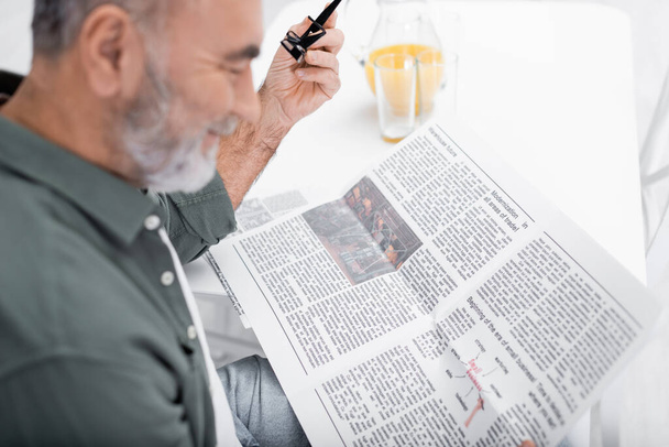 θολή ανώτερος άνθρωπος χαμογελά ενώ διαβάζετε εφημερίδα κοντά χυμό πορτοκαλιού στην κουζίνα - Φωτογραφία, εικόνα