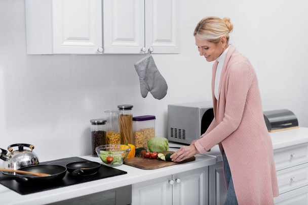 casalinga sorridente che taglia il cetriolo mentre prepara l'insalata in cucina - Foto, immagini