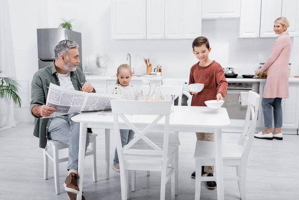 старший мужчина с газетой глядя на внучку, держащую чашки на кухне - Фото, изображение