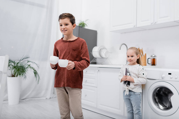 smiling kids holding cups and cutlery in kitchen - Foto, Bild