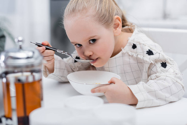 дівчина снідає на кухні біля розмитого чайника і чашок
 - Фото, зображення
