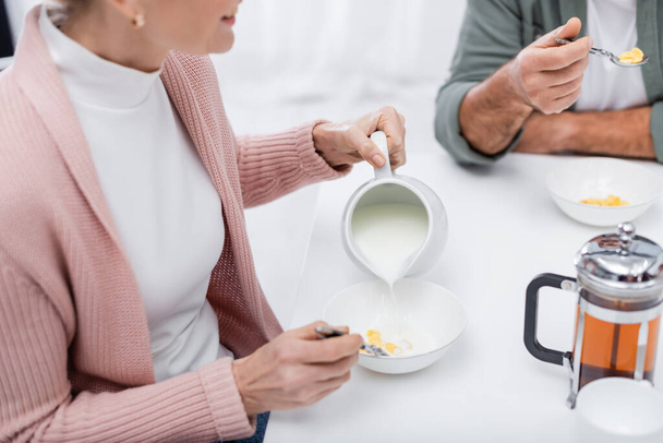 abgeschnittene Ansicht einer Frau, die beim Frühstück mit ihrem Mann Milch in eine Schüssel mit Cornflakes gießt - Foto, Bild