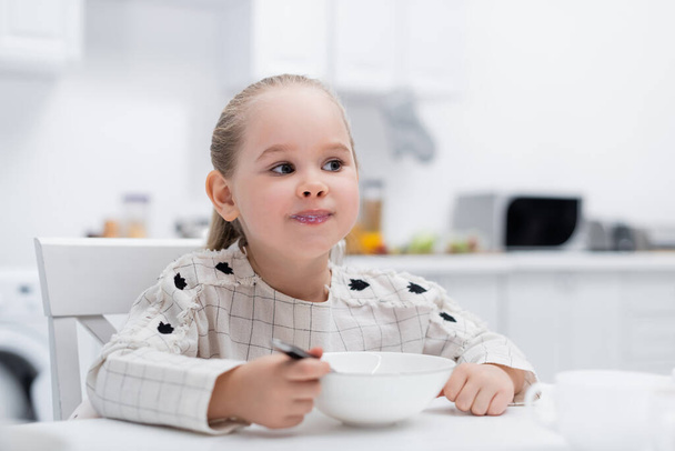 little girl eating breakfast while sitting in blurred kitchen - Fotó, kép