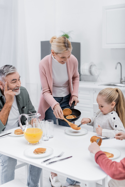 smiling woman holding frying pan with pancakes near husband and grandchildren - Foto, Bild