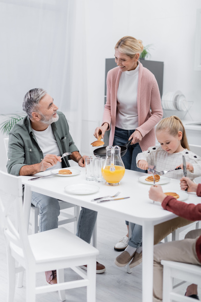 Женщина средних лет держит сковородку с блинами рядом с счастливым мужем и внуками - Фото, изображение
