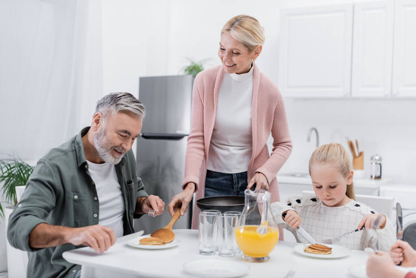 happy woman serving pancakes for husband during breakfast with grandchildren - Foto, imagen