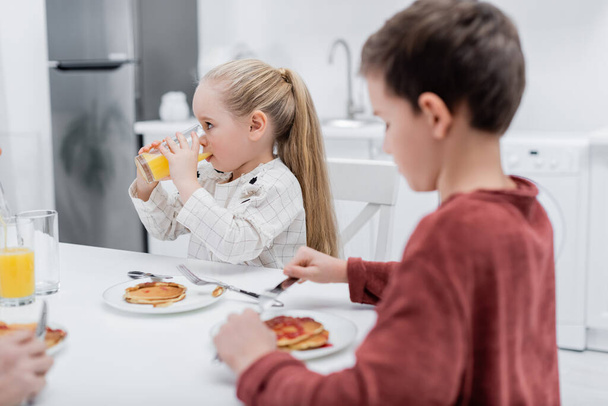 girl drinking orange juice near pancakes and blurred brother - Fotó, kép