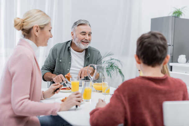 весёлый пожилой мужчина завтракает с размытыми внуками и женой на кухне - Фото, изображение