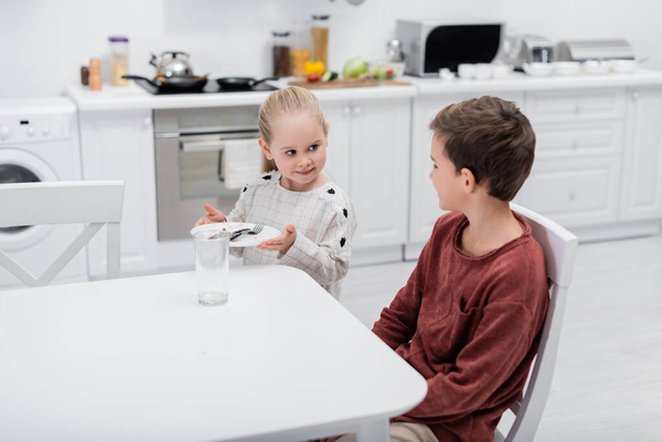 улыбающаяся девушка держит тарелку со столовыми приборами рядом с братом, сидящим за кухонным столом - Фото, изображение