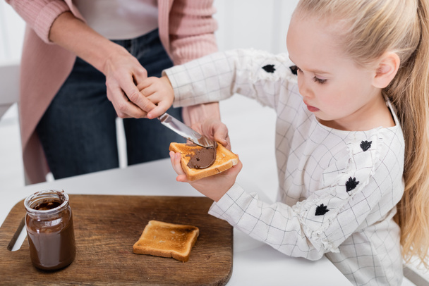 a kislány csokipürét terjeszt a kenyérre, miközben szendvicset készít a nagyival. - Fotó, kép