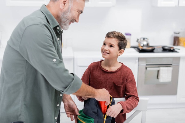 smiling senior man packing backpack of grandson in kitchen - Foto, immagini