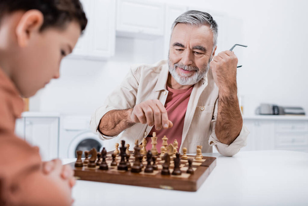 pleased senior man playing chess with blurred grandson in kitchen - Fotó, kép