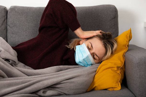 Young woman with coronavirus is having headache and fever, lying on the sofa wrapped in a balnket - Valokuva, kuva