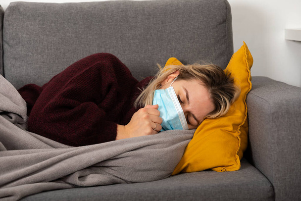 Young woman with coronavirus is having headache and fever, lying on the sofa wrapped in a balnket - Foto, imagen